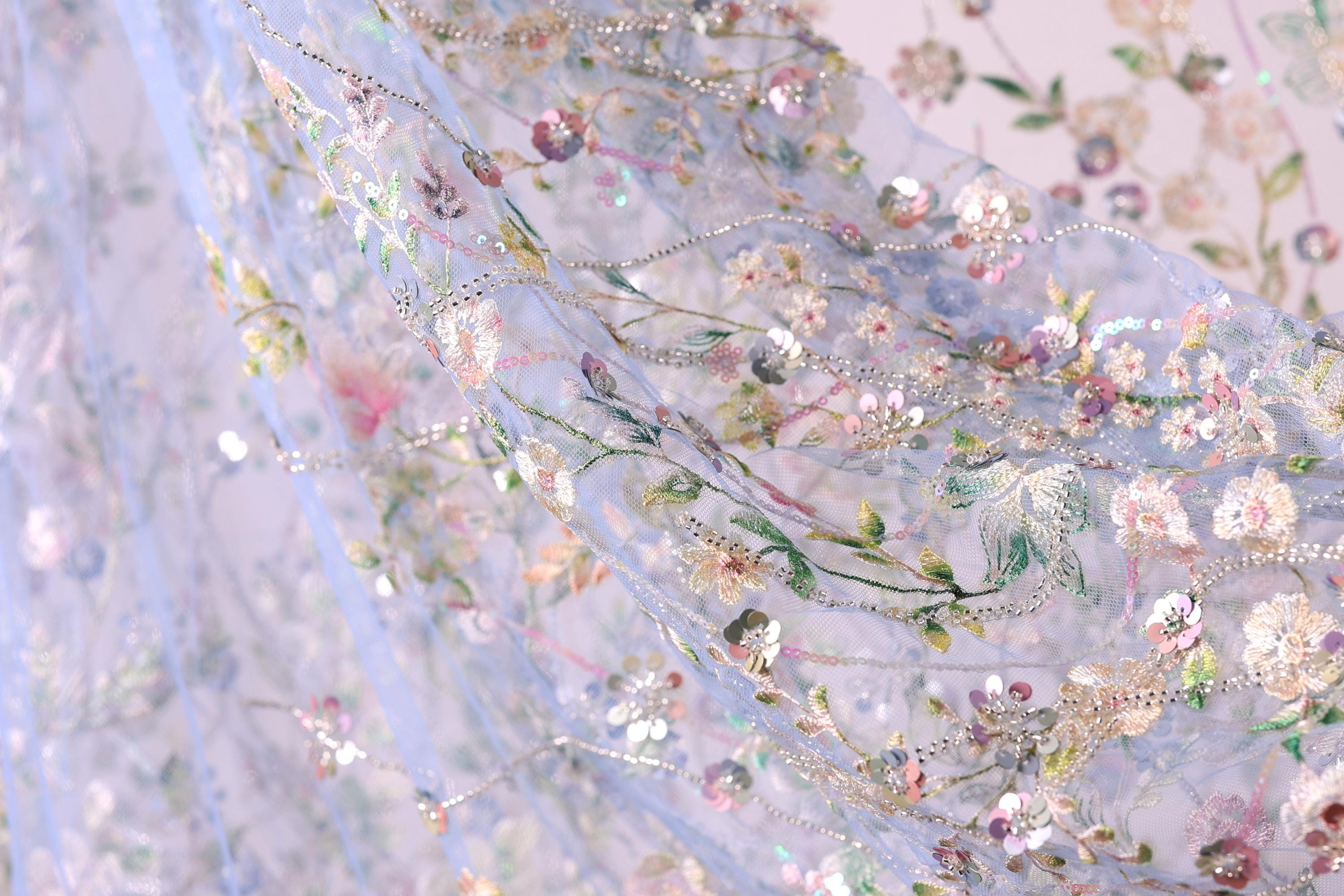 The image is a close-up of a delicate, intricately embroidered fabric with a floral pattern. The fabric appears to be made of sheer material, giving it a soft and delicate appearance. The embroidery features a variety of flowers and leaves, creating a visually appealing and intricate design. The colors used in the embroidery are predominantly pink, white, and green, which contribute to the overall beauty of the fabric.