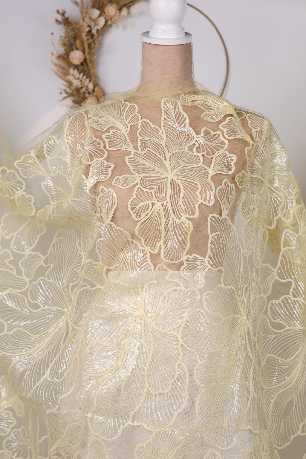 a mannequin wearing a white dress with a yellow floral pattern. The dress has a sheer, lace-like design with a delicate, flower-like pattern. The mannequin is positioned in front of a wreath, which adds a decorative element to the scene.