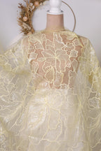 a mannequin wearing a white dress with a yellow floral pattern. The dress has a sheer, lace-like design with a delicate, flower-like pattern. The mannequin is positioned in front of a wreath, which adds a decorative element to the scene.