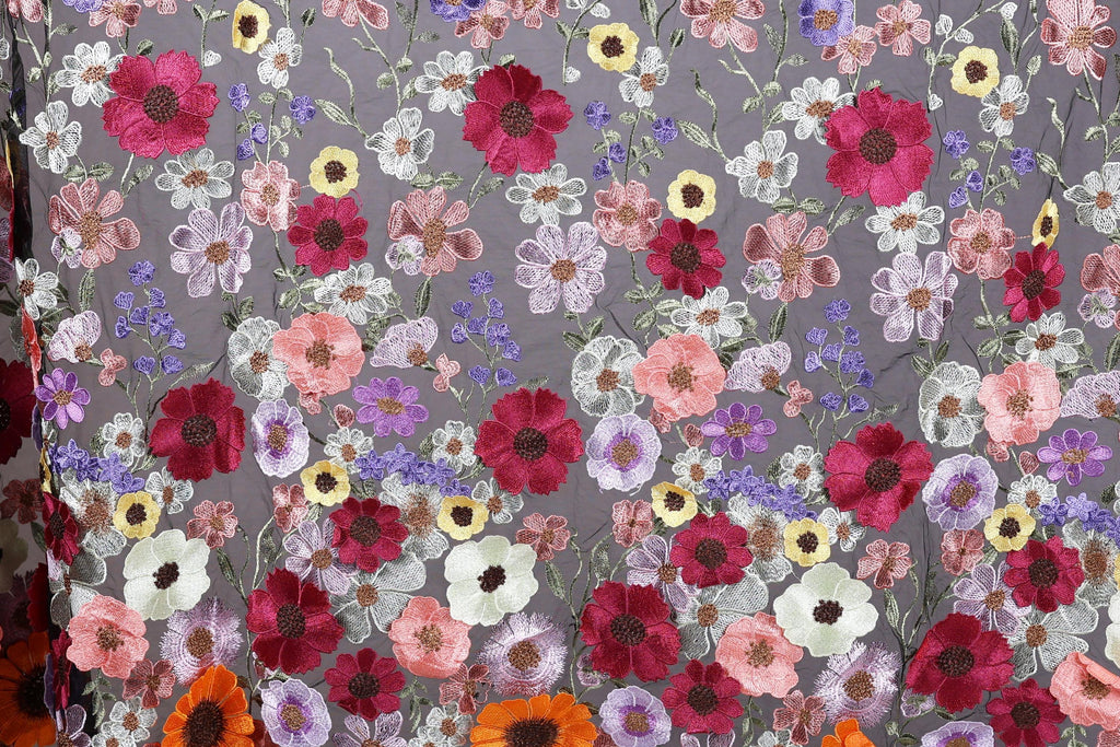 a close-up view of a fabric with a vibrant floral pattern. The fabric is adorned with a variety of colorful flowers, including red, pink, white, and yellow flowers, as well as purple and orange flowers. The flowers are intricately embroidered or sewn onto the fabric, creating a visually striking and detailed design. The image does not contain any text or other discernible objects, and the focus is solely on the floral pattern on the fabric.