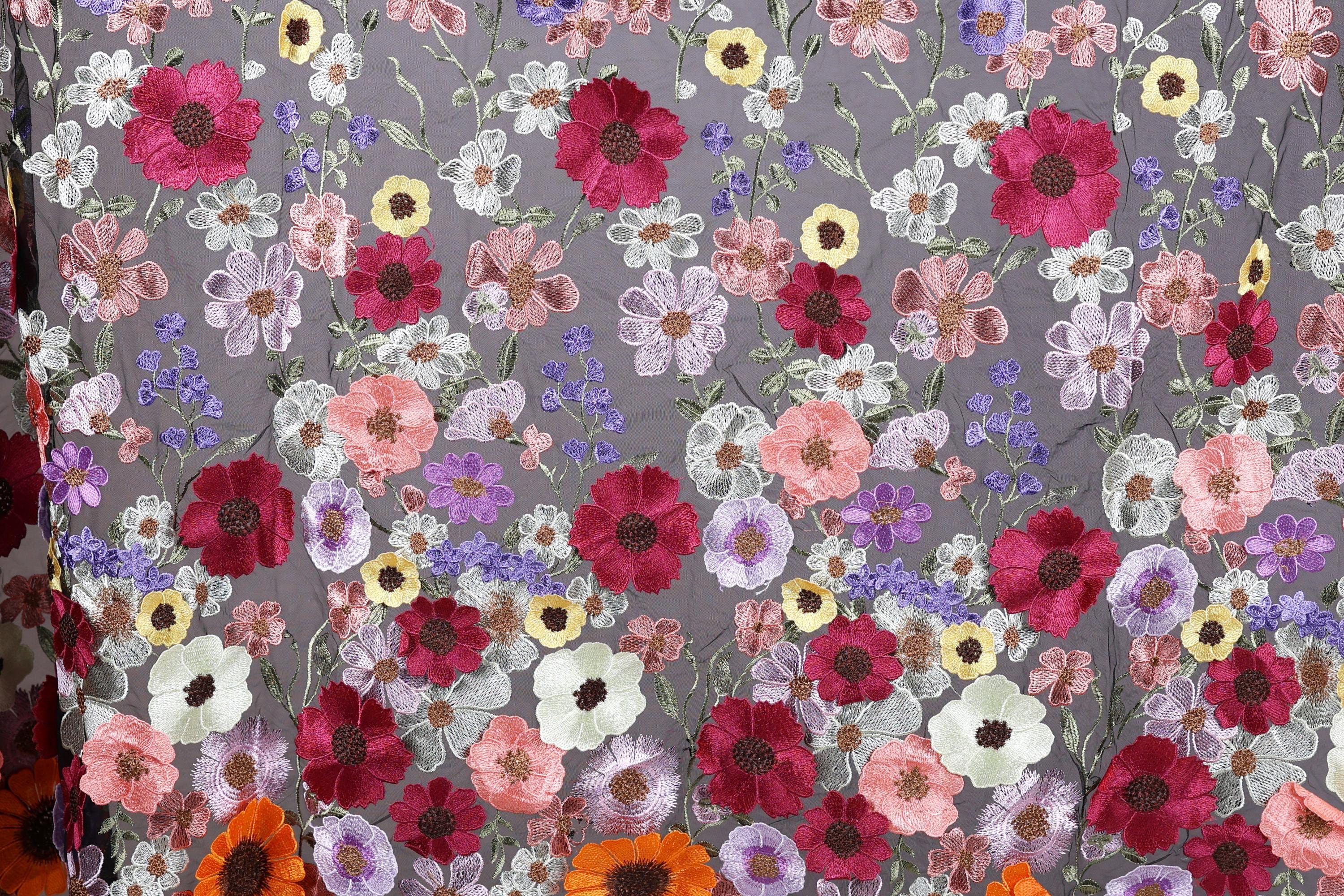 a close-up view of a fabric with a vibrant floral pattern. The fabric is adorned with a variety of colorful flowers, including red, pink, white, and yellow flowers, as well as purple and orange flowers. The flowers are intricately embroidered or sewn onto the fabric, creating a visually striking and detailed design. The image does not contain any text or other discernible objects, and the focus is solely on the floral pattern on the fabric.