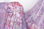a close-up view of a fabric with a floral pattern in shades of purple and pink, with some white and green accents. The fabric appears to be made of sequins or other embellishments, creating a visually striking and intricate design.