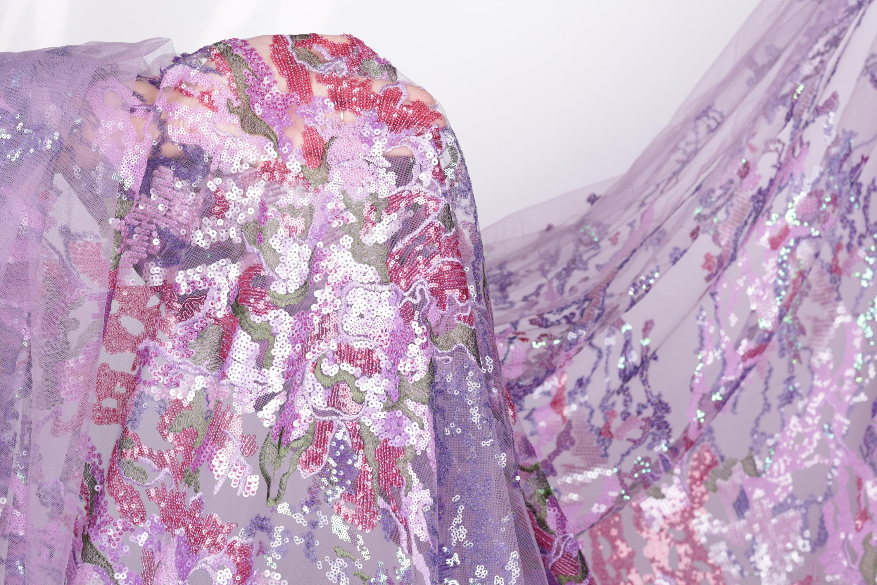a close-up view of a fabric with a floral pattern in shades of purple and pink, with some white and green accents. The fabric appears to be made of sequins or other embellishments, creating a visually striking and intricate design.