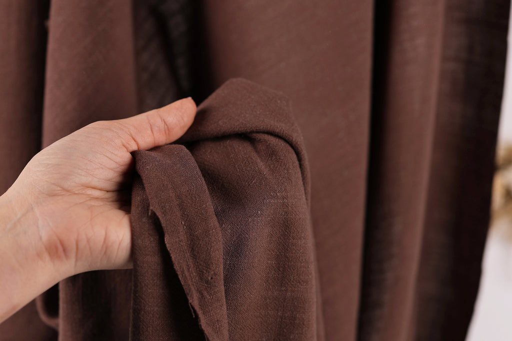 A hand is holding a brown fabric, possibly a piece of clothing or a textile, in front of a brown curtain.