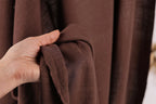 A hand is holding a brown fabric, possibly a piece of clothing or a textile, in front of a brown curtain.