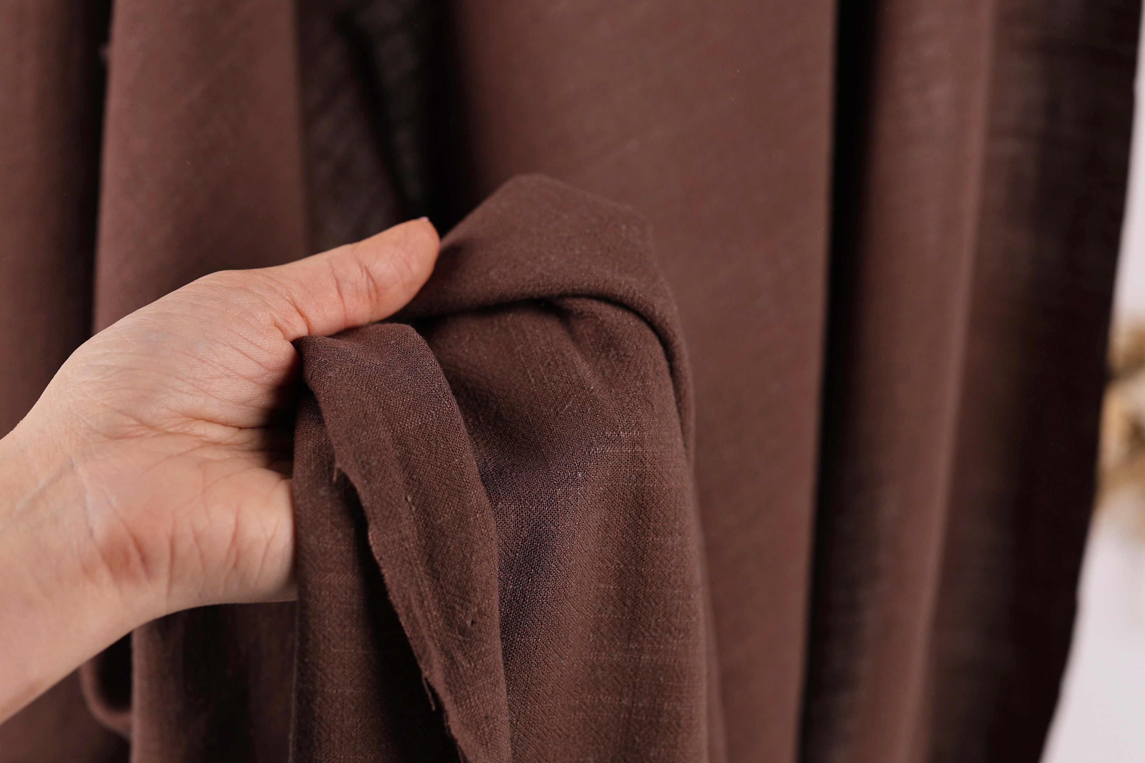 A hand is holding a brown fabric, possibly a piece of clothing or a textile, in front of a brown curtain.