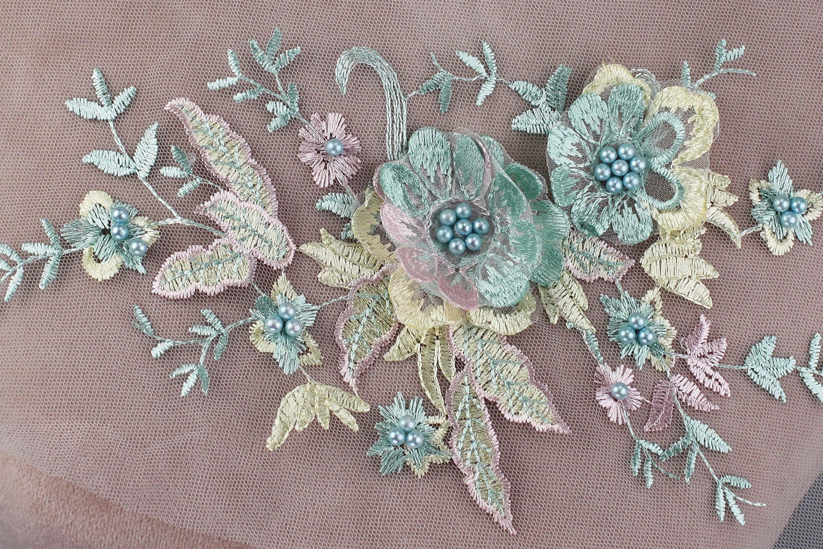 a close-up view of a delicate embroidered floral design on a fabric surface. The design includes various flowers and leaves, with the flowers being the main focus. The flowers are intricately crafted with beads, adding a touch of elegance and detail to the overall design. The arrangement of the flowers and leaves creates a visually appealing and harmonious composition.