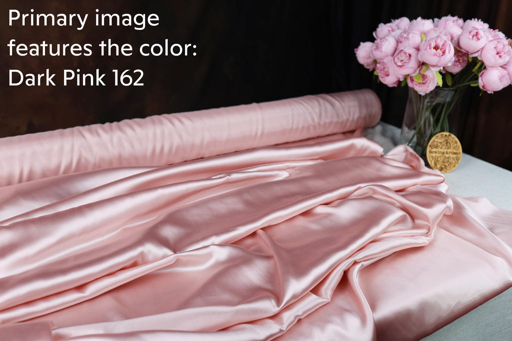 a roll of pink satin fabric, a vase of pink flowers, and a gold medallion on a table.