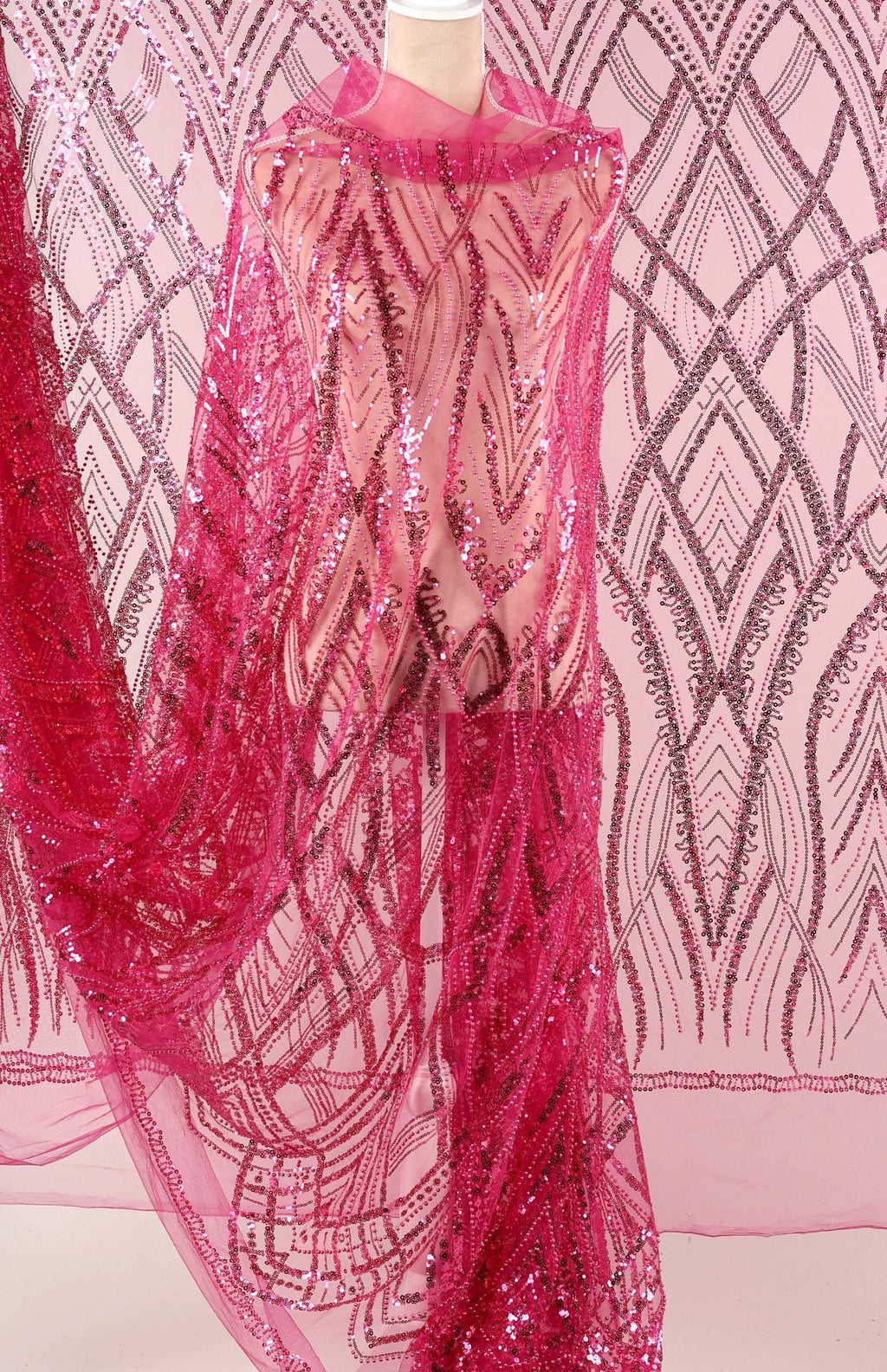a mannequin wearing a pink sequin dress with a sheer overlay. The dress has a long, flowing skirt and is adorned with a pattern of pink sequins. The background is a pink and white patterned fabric, which complements the overall color scheme of the dress.