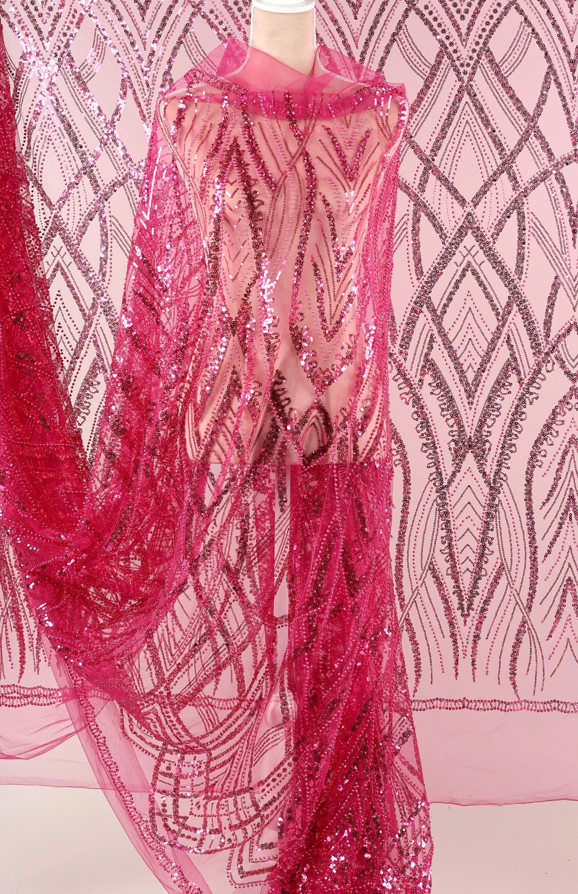 a mannequin wearing a pink sequin dress with a sheer overlay. The dress has a long, flowing skirt and is adorned with a pattern of pink sequins. The background is a pink and white patterned fabric, which complements the overall color scheme of the dress.