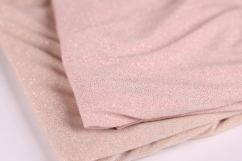 a close-up view of a fabric with a metallic, glittery texture. The fabric appears to be a light pink color, and it is folded or crumpled, creating a sense of depth and texture.