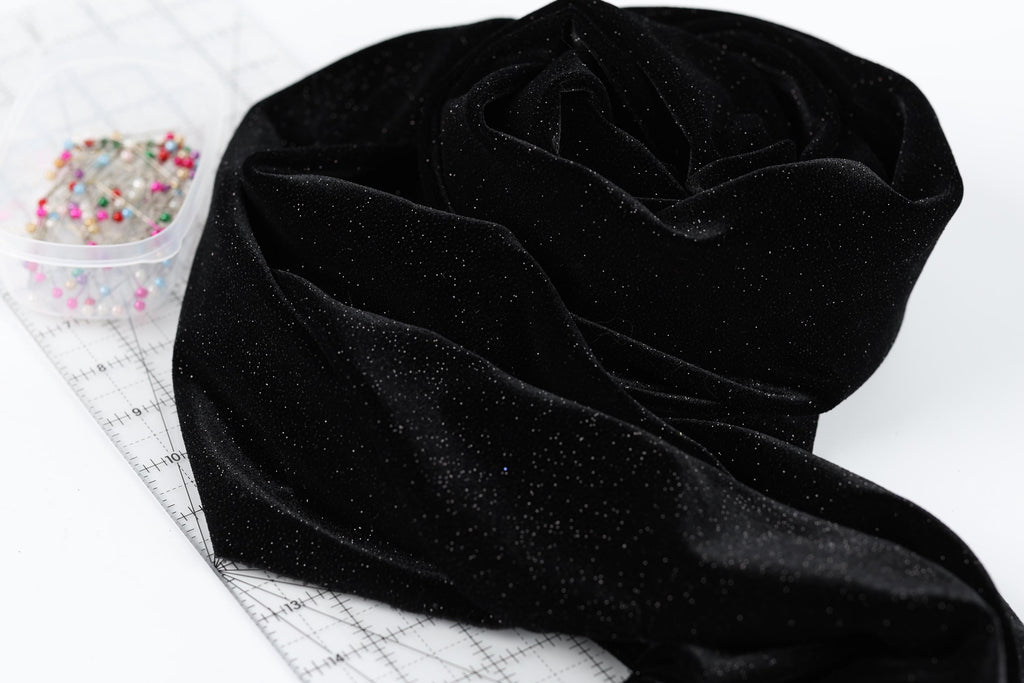 a black velvet fabric with a glittery texture, placed on a white surface next to a clear plastic container filled with colorful pins.