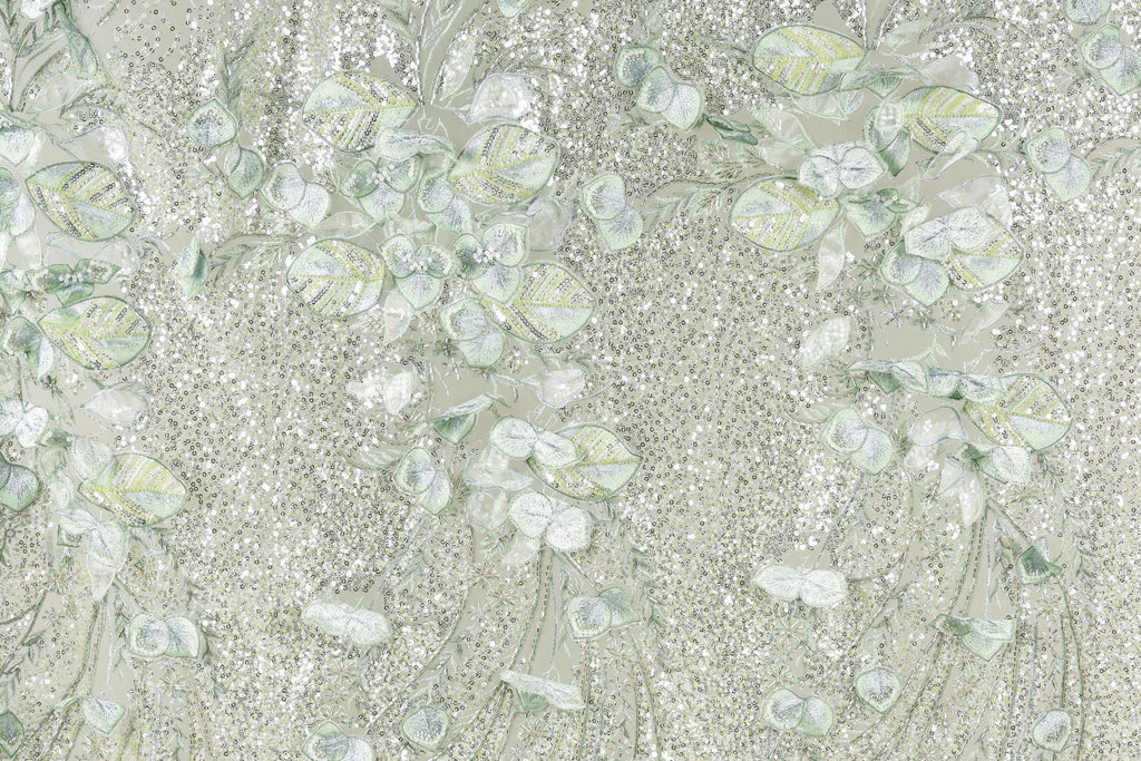 a close-up view of a fabric with a floral pattern, including leaves and flowers in shades of green and white. The fabric appears to be made of a light-colored material, possibly lace or a similar type of textile. The pattern is intricate and detailed, with the leaves and flowers appearing to be hand-stitched or embroidered onto the fabric.