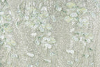 a close-up view of a fabric with a floral pattern, including leaves and flowers in shades of green and white. The fabric appears to be made of a light-colored material, possibly lace or a similar type of textile. The pattern is intricate and detailed, with the leaves and flowers appearing to be hand-stitched or embroidered onto the fabric.