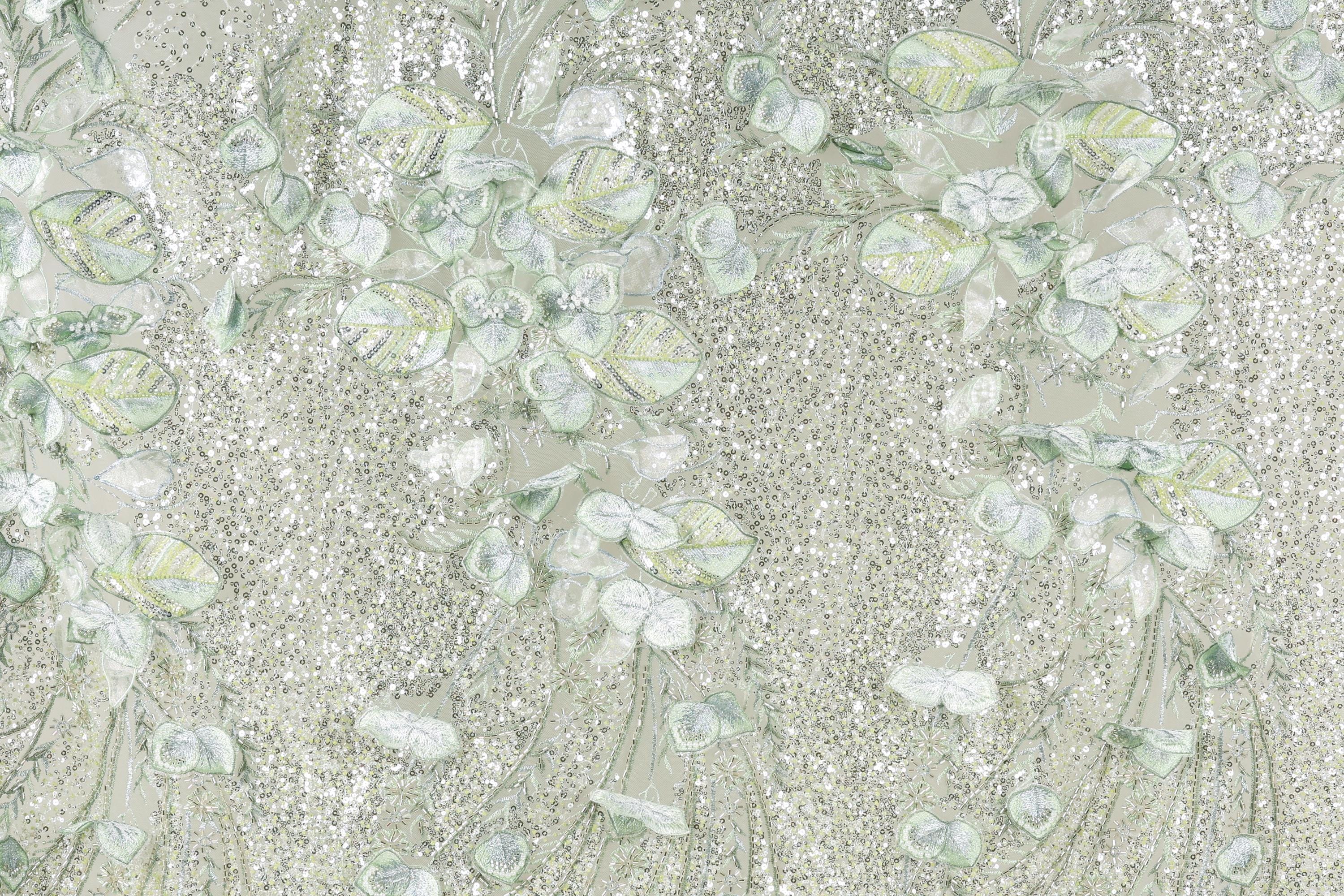 a close-up view of a fabric with a floral pattern, including leaves and flowers in shades of green and white. The fabric appears to be made of a light-colored material, possibly lace or a similar type of textile. The pattern is intricate and detailed, with the leaves and flowers appearing to be hand-stitched or embroidered onto the fabric.