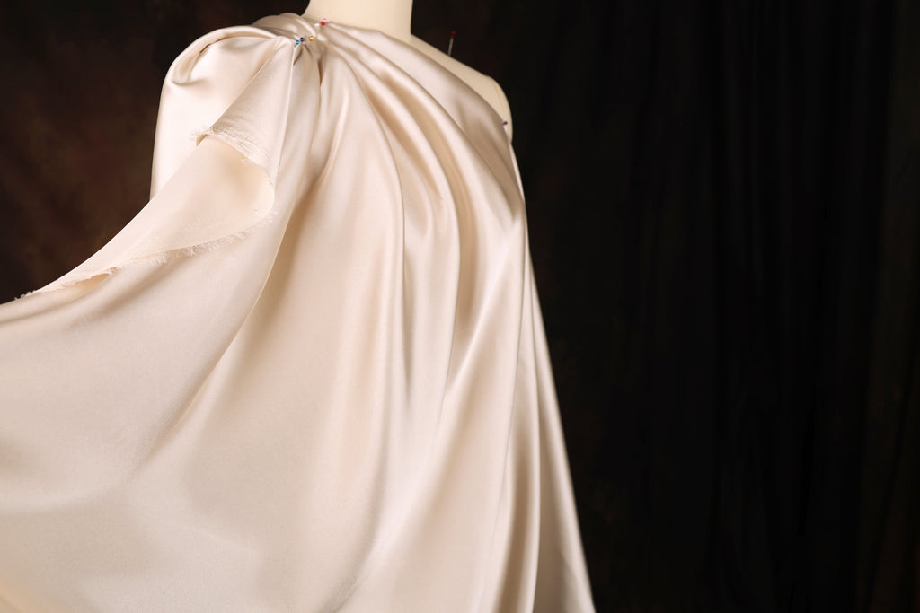 A flowing, draped fabric in a pale color, possibly white or light pink, is displayed against a dark background.