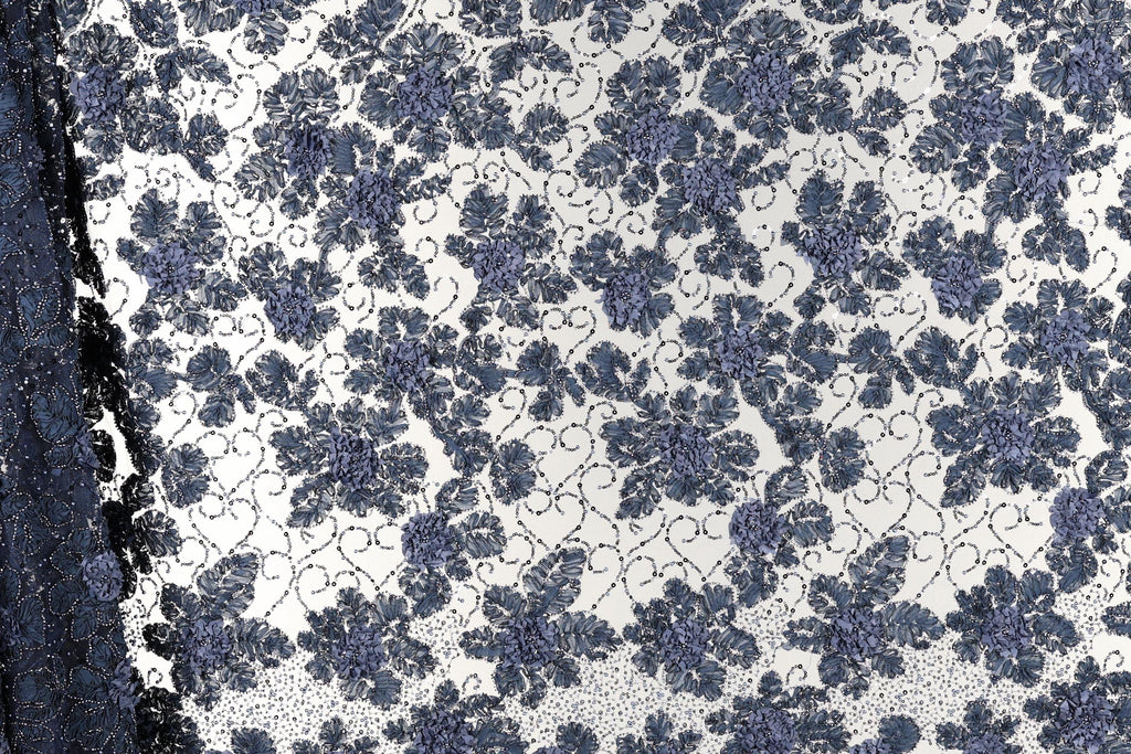 a close-up view of a fabric with a floral pattern in shades of blue and white. The pattern consists of numerous small blue flowers arranged in a repeating design across the fabric's surface. The fabric appears to be a lace-like material, with the intricate floral pattern adding a decorative element to the design.