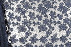a close-up view of a fabric with a floral pattern in shades of blue and white. The pattern consists of numerous small blue flowers arranged in a repeating design across the fabric's surface. The fabric appears to be a lace-like material, with the intricate floral pattern adding a decorative element to the design.
