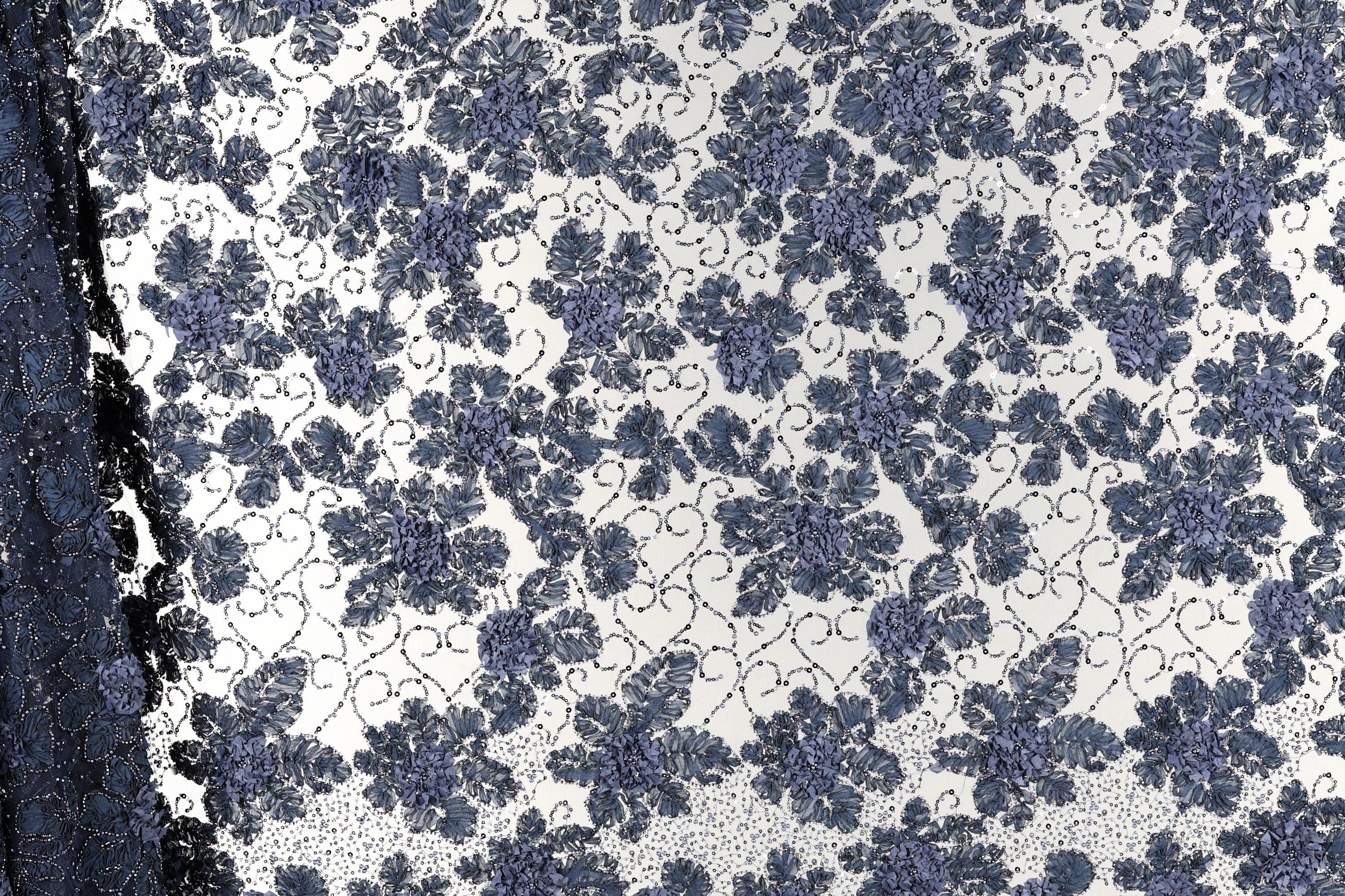 a close-up view of a fabric with a floral pattern in shades of blue and white. The pattern consists of numerous small blue flowers arranged in a repeating design across the fabric's surface. The fabric appears to be a lace-like material, with the intricate floral pattern adding a decorative element to the design.