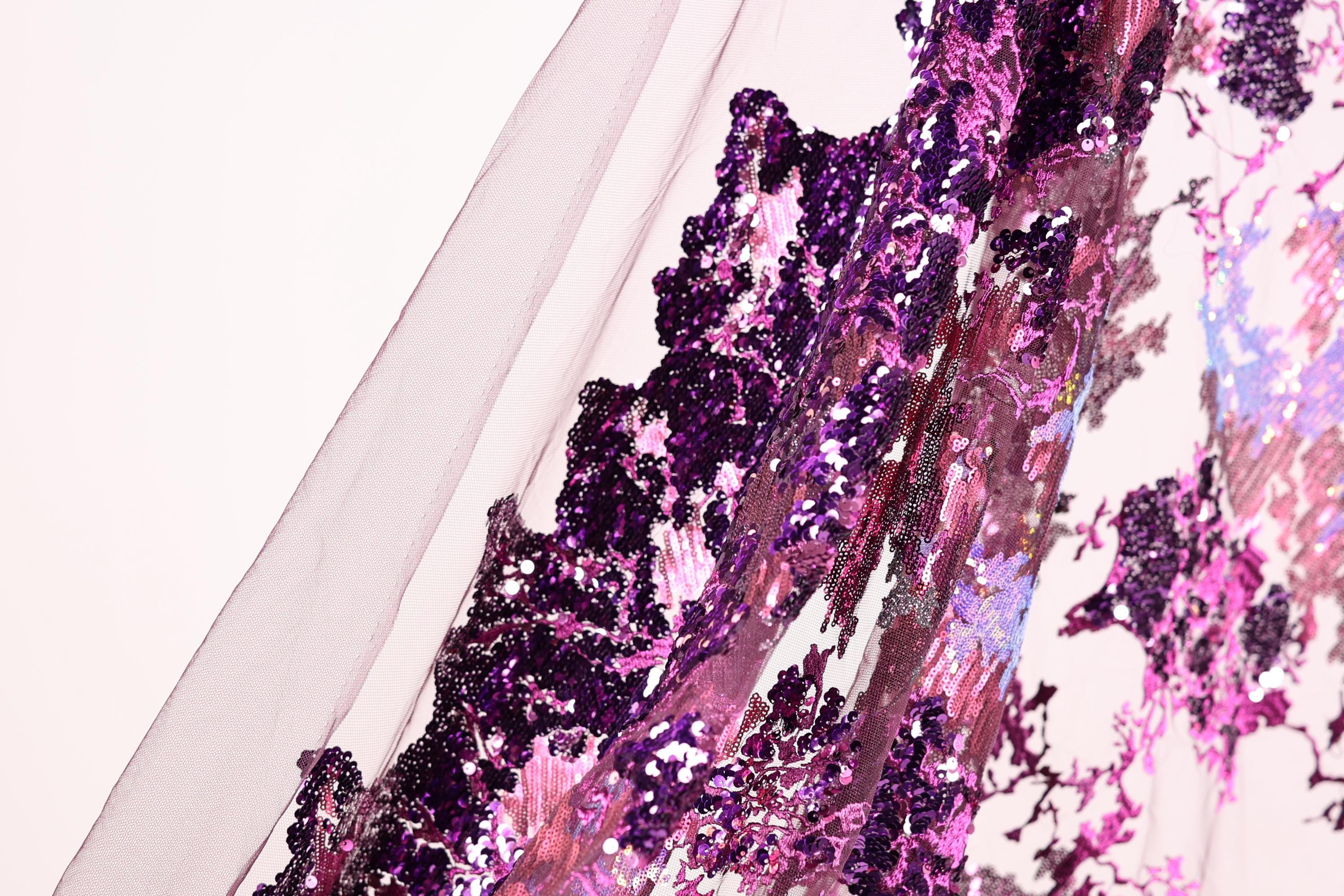 a close-up view of a fabric with a vibrant purple and pink sequin pattern. The fabric appears to be a sheer material, with a mix of purple and pink sequins that create a visually striking and intricate design.