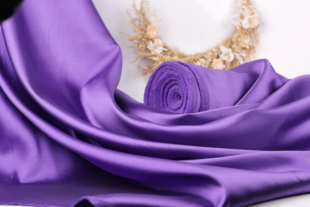a close-up view of a purple satin fabric, with a rolled-up piece of the fabric visible in the background.