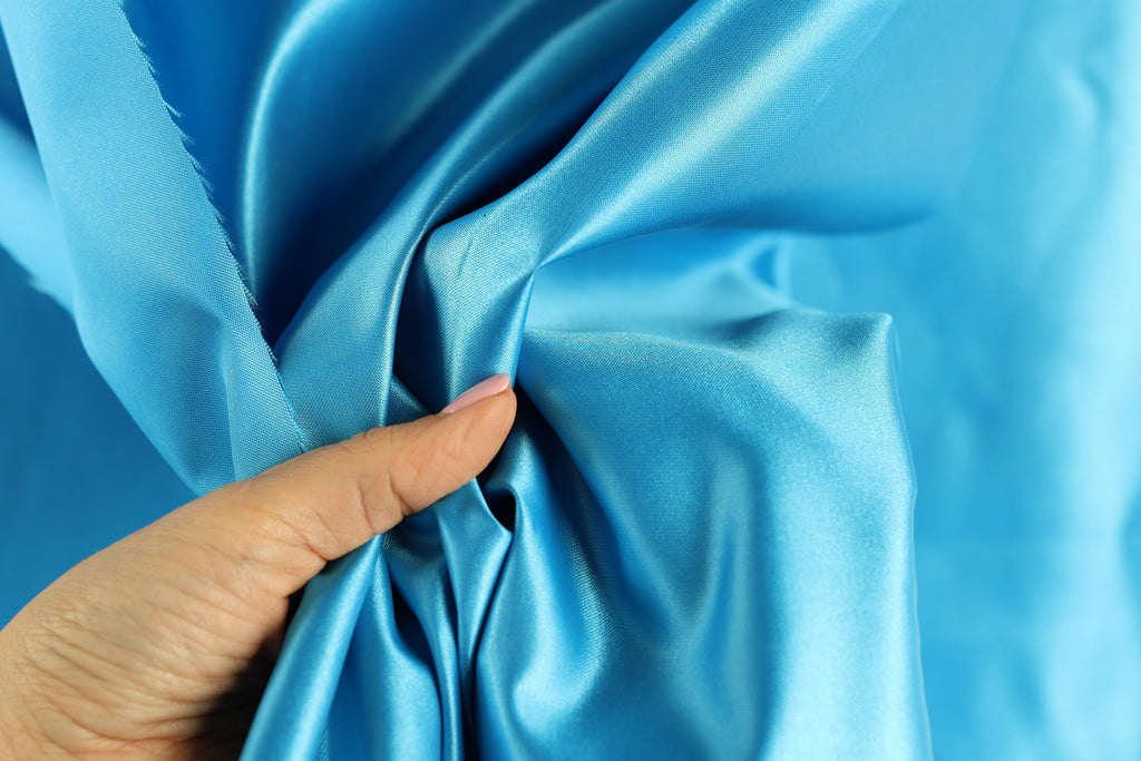 A hand is holding a piece of blue satin fabric, with the fabric being crumpled and wrinkled.