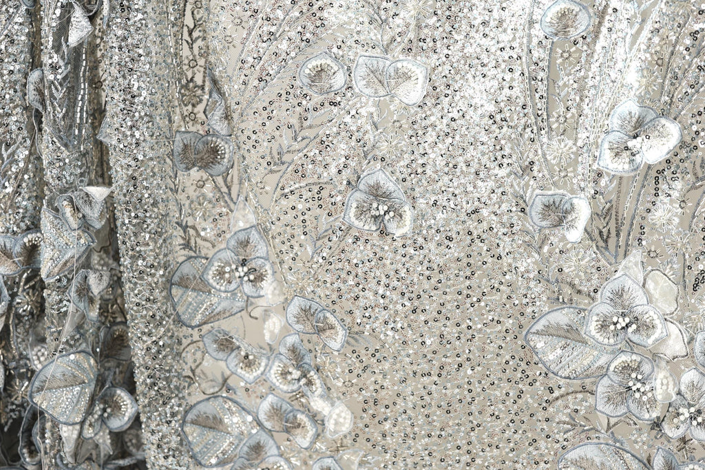 The image is a close-up view of a fabric with a floral pattern, featuring a mix of silver and white sequins and leaves. The fabric appears to be made of a shiny, metallic material, giving it a metallic, glittery appearance.
