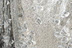 The image is a close-up view of a fabric with a floral pattern, featuring a mix of silver and white sequins and leaves. The fabric appears to be made of a shiny, metallic material, giving it a metallic, glittery appearance.