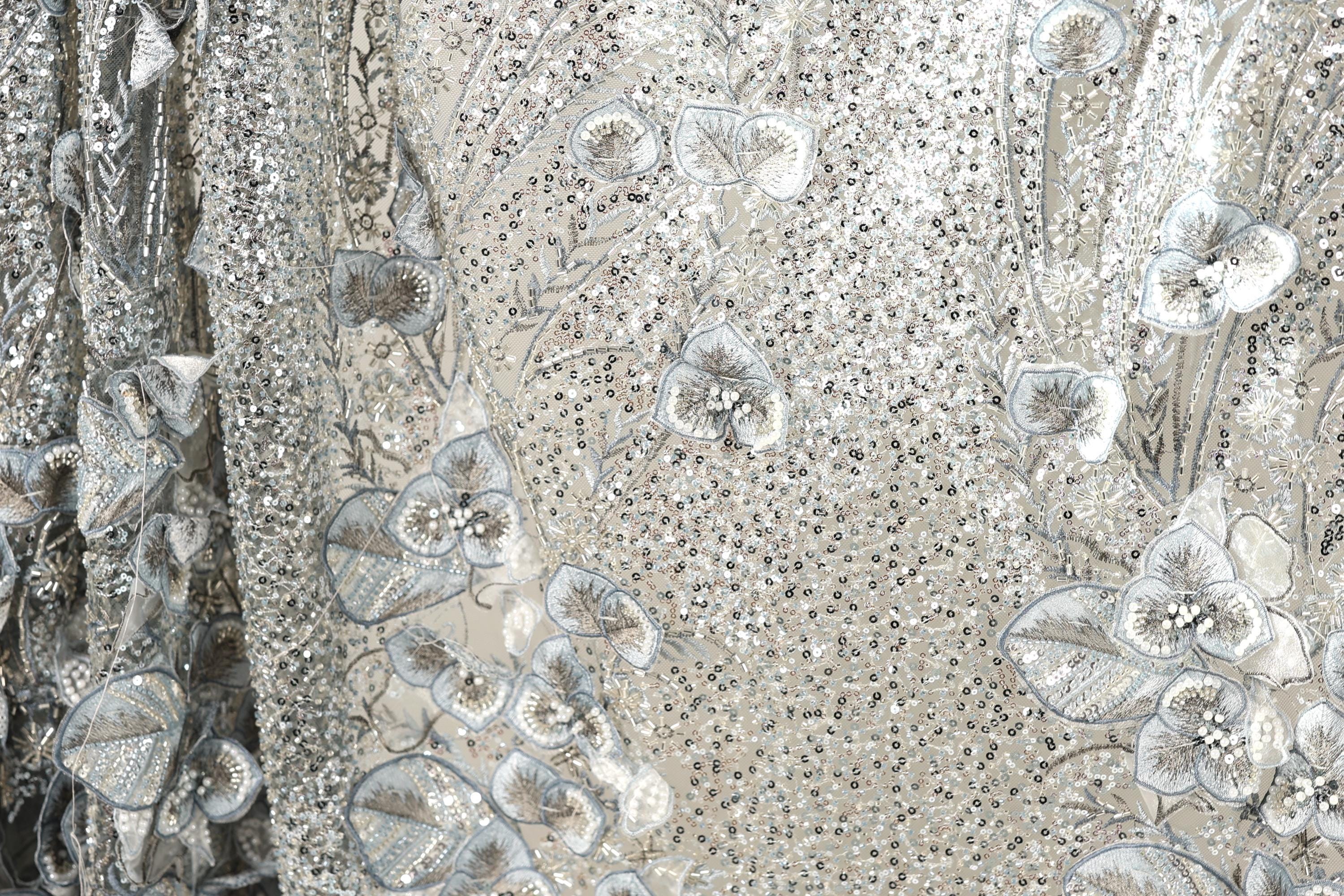 The image is a close-up view of a fabric with a floral pattern, featuring a mix of silver and white sequins and leaves. The fabric appears to be made of a shiny, metallic material, giving it a metallic, glittery appearance.