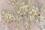 a close-up view of a delicate, intricate piece of embroidered lace or fabric adorned with gold and white floral elements. The design is composed of multiple flowers and leaves, creating a visually appealing and detailed pattern.