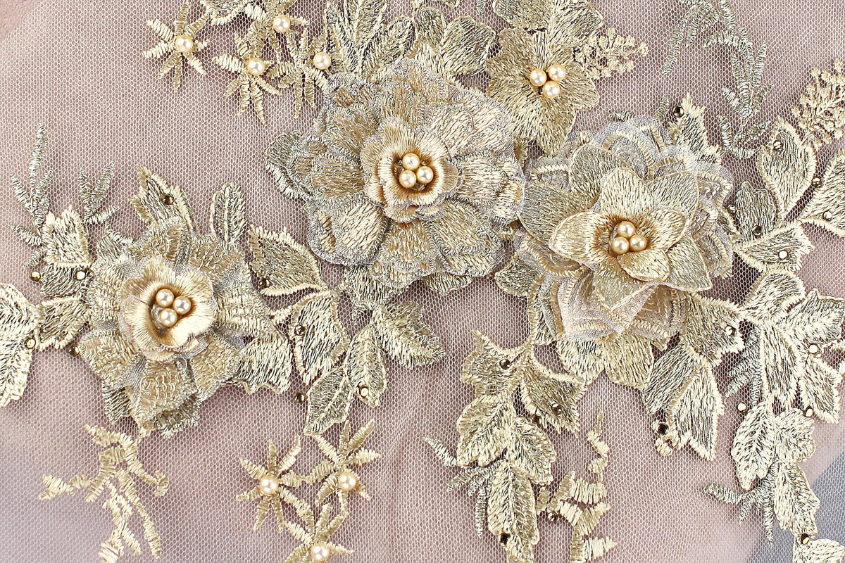 a close-up view of a delicate, intricate piece of embroidered lace or fabric adorned with gold and white floral elements. The design is composed of multiple flowers and leaves, creating a visually appealing and detailed pattern.
