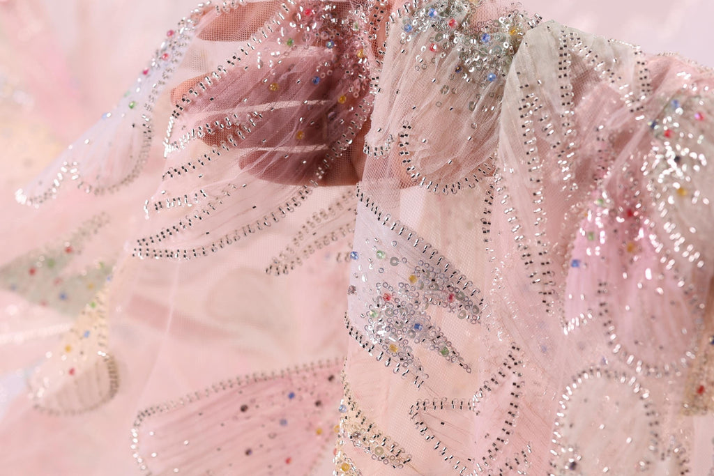 The image is a close-up of a delicate, intricately designed fabric with a pink and white color scheme, adorned with sparkling embellishments.