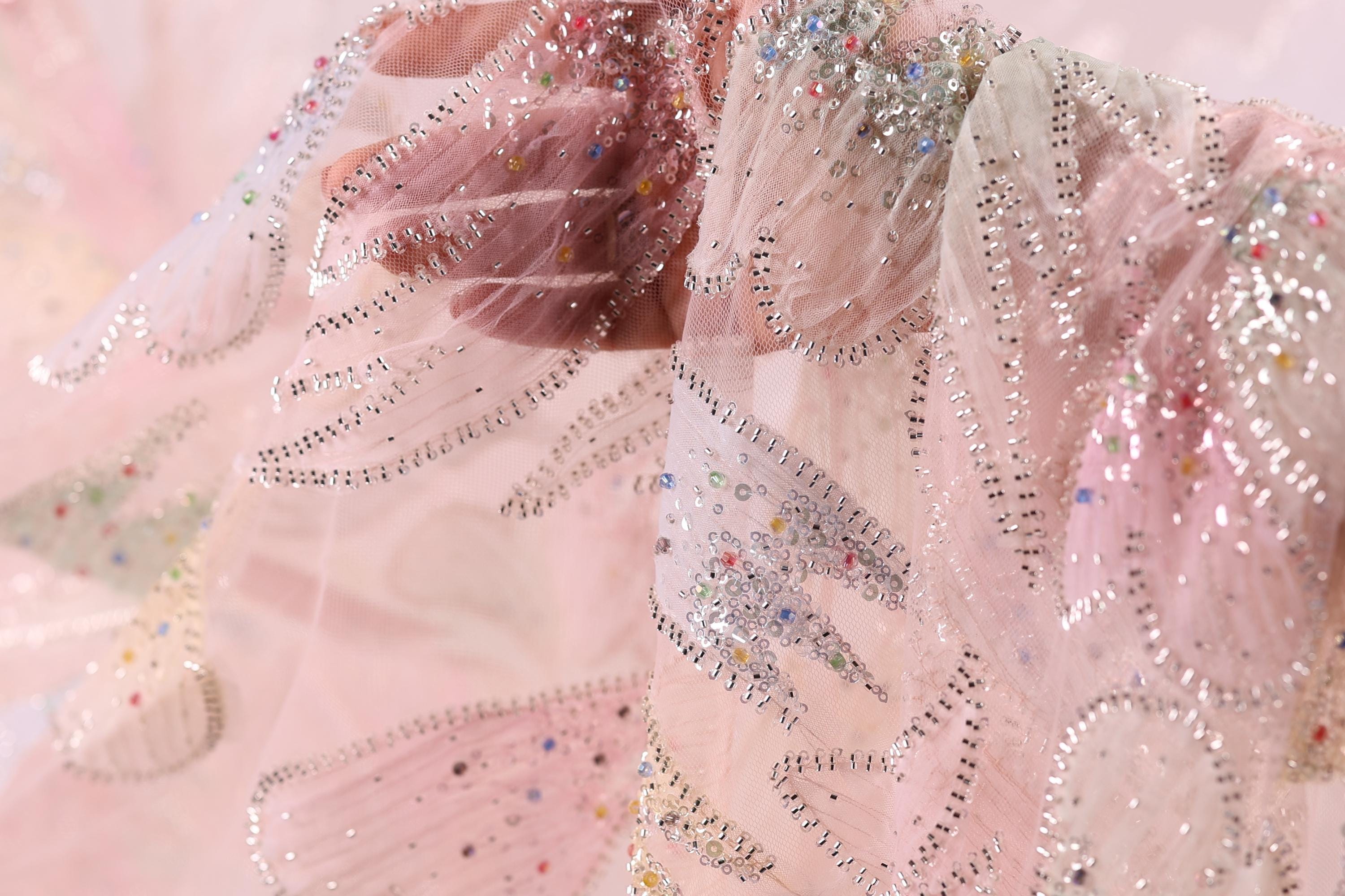 The image is a close-up of a delicate, intricately designed fabric with a pink and white color scheme, adorned with sparkling embellishments.