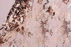 a close-up view of a fabric with a metallic, sequined texture. The fabric appears to be a light pink color, with a pattern of small, irregularly shaped sequins or glittering particles covering the surface. The fabric is folded or crumpled, creating a sense of depth and texture in the image.