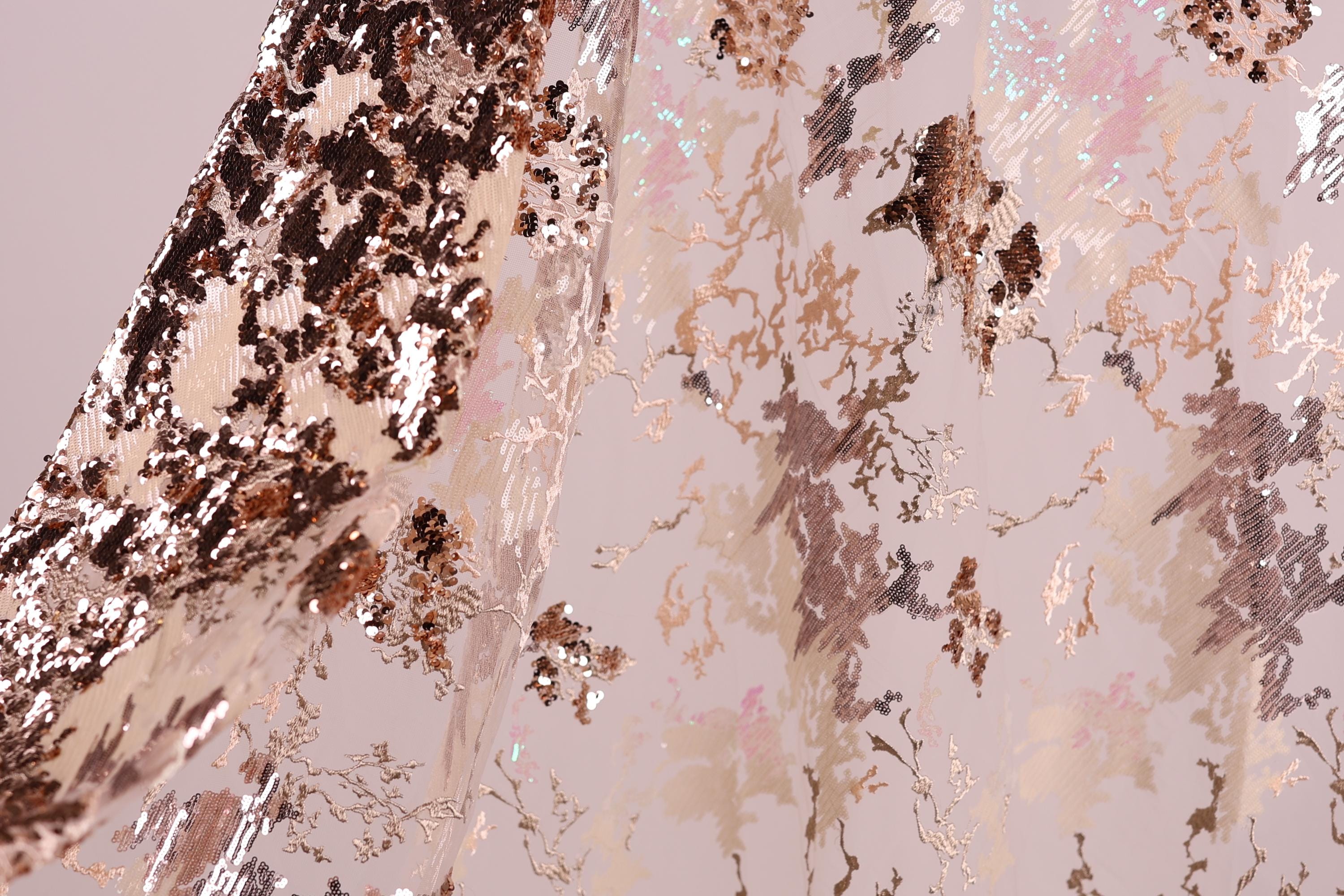 a close-up view of a fabric with a metallic, sequined texture. The fabric appears to be a light pink color, with a pattern of small, irregularly shaped sequins or glittering particles covering the surface. The fabric is folded or crumpled, creating a sense of depth and texture in the image.