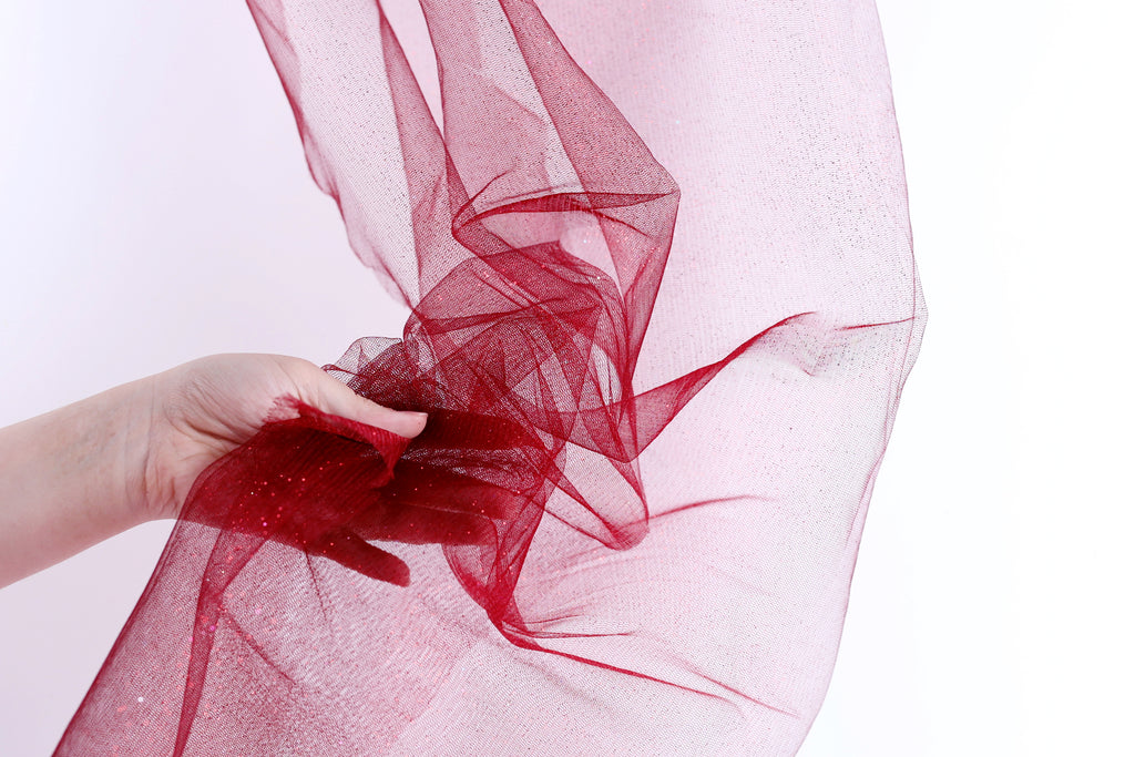 A hand is holding a piece of red tulle fabric, which is being twisted and draped over a white background.