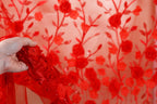 A hand is holding a piece of red fabric with a floral pattern, against a background of red fabric with similar floral patterns.