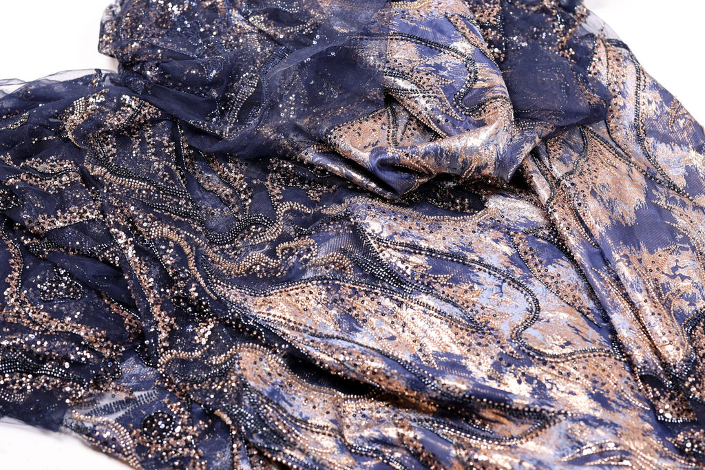 a close-up view of a fabric with a blue and gold pattern, featuring a mix of metallic and organic elements. The fabric appears to be a blend of lace and sequin, with the lace having a darker blue hue and the sequins adding a touch of gold. The fabric is crumpled and wrinkled, giving it a textured and layered appearance.