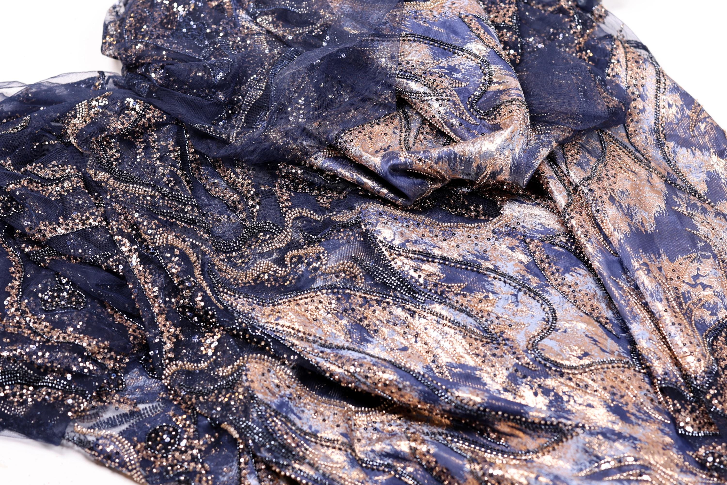 a close-up view of a fabric with a blue and gold pattern, featuring a mix of metallic and organic elements. The fabric appears to be a blend of lace and sequin, with the lace having a darker blue hue and the sequins adding a touch of gold. The fabric is crumpled and wrinkled, giving it a textured and layered appearance.
