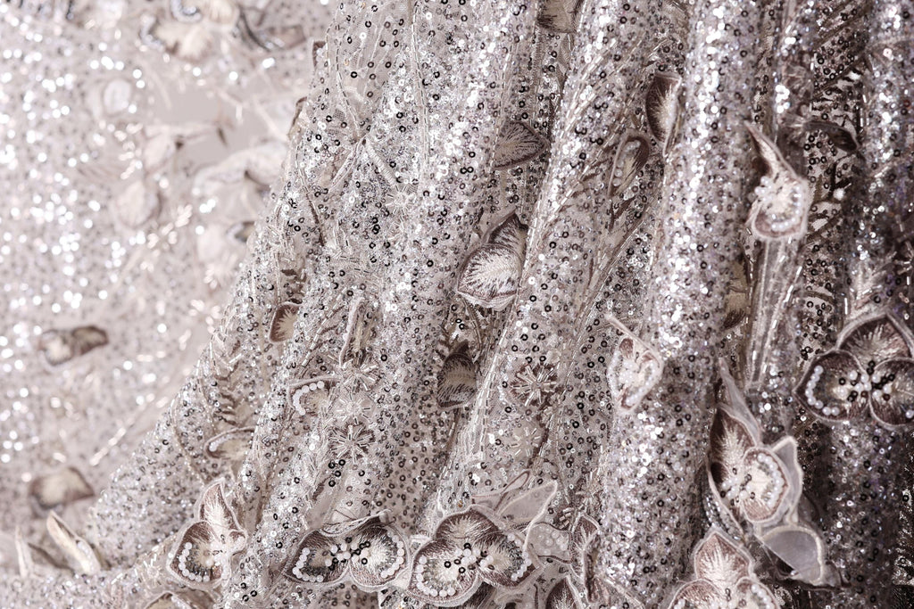 The image is a close-up of a fabric with a silver sequin pattern, featuring a delicate floral design.