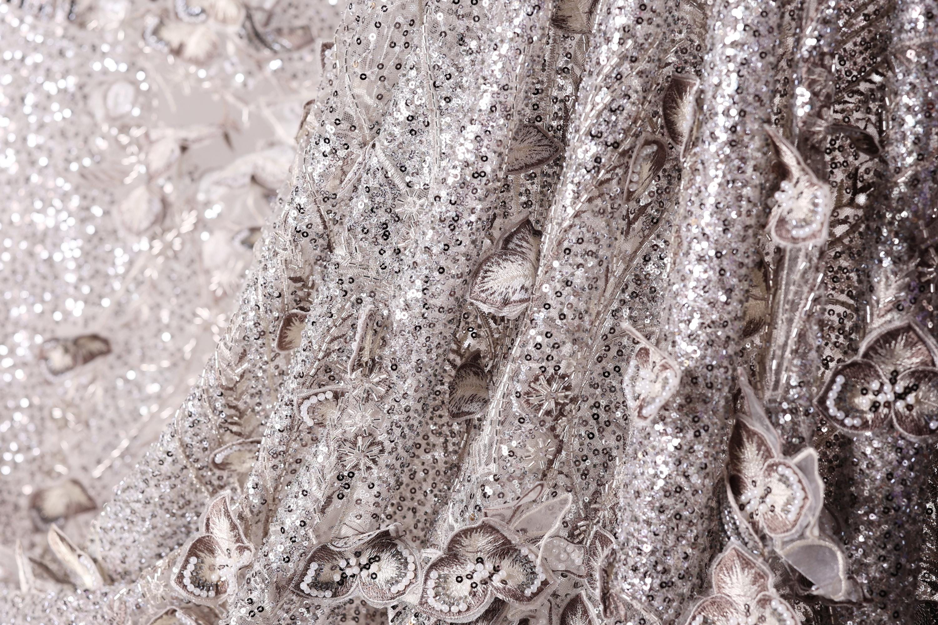 The image is a close-up of a fabric with a silver sequin pattern, featuring a delicate floral design.