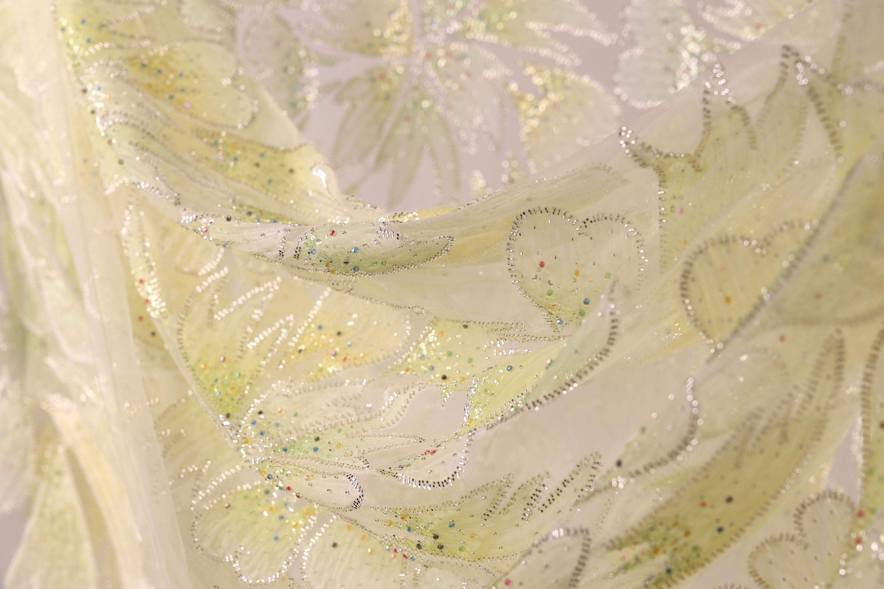 The image is a close-up of a fabric with a floral pattern, featuring yellow and green colors and small, multicolored beads or sequins.