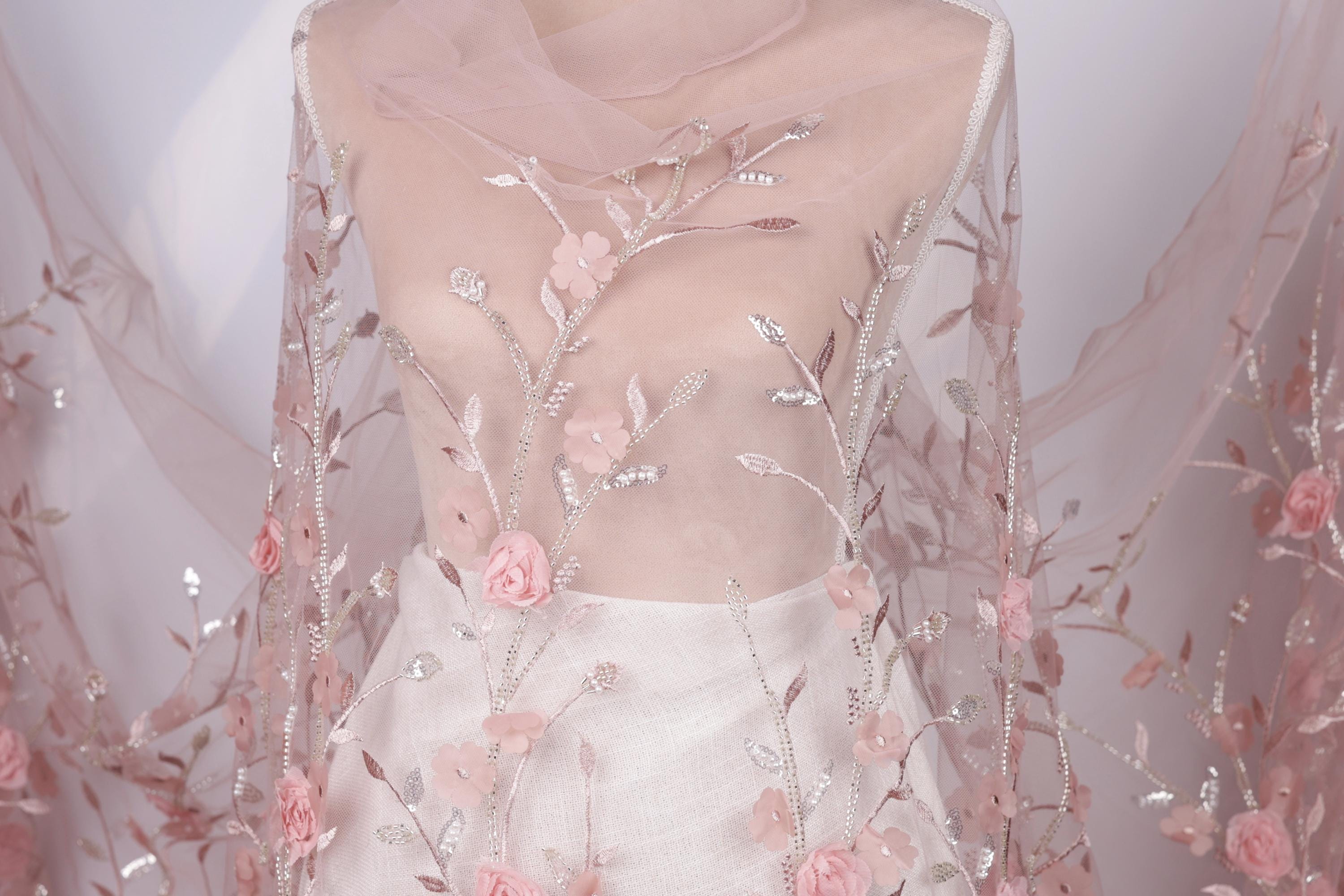 A mannequin wearing a white dress with pink floral embroidery and a sheer pink fabric draped over it.