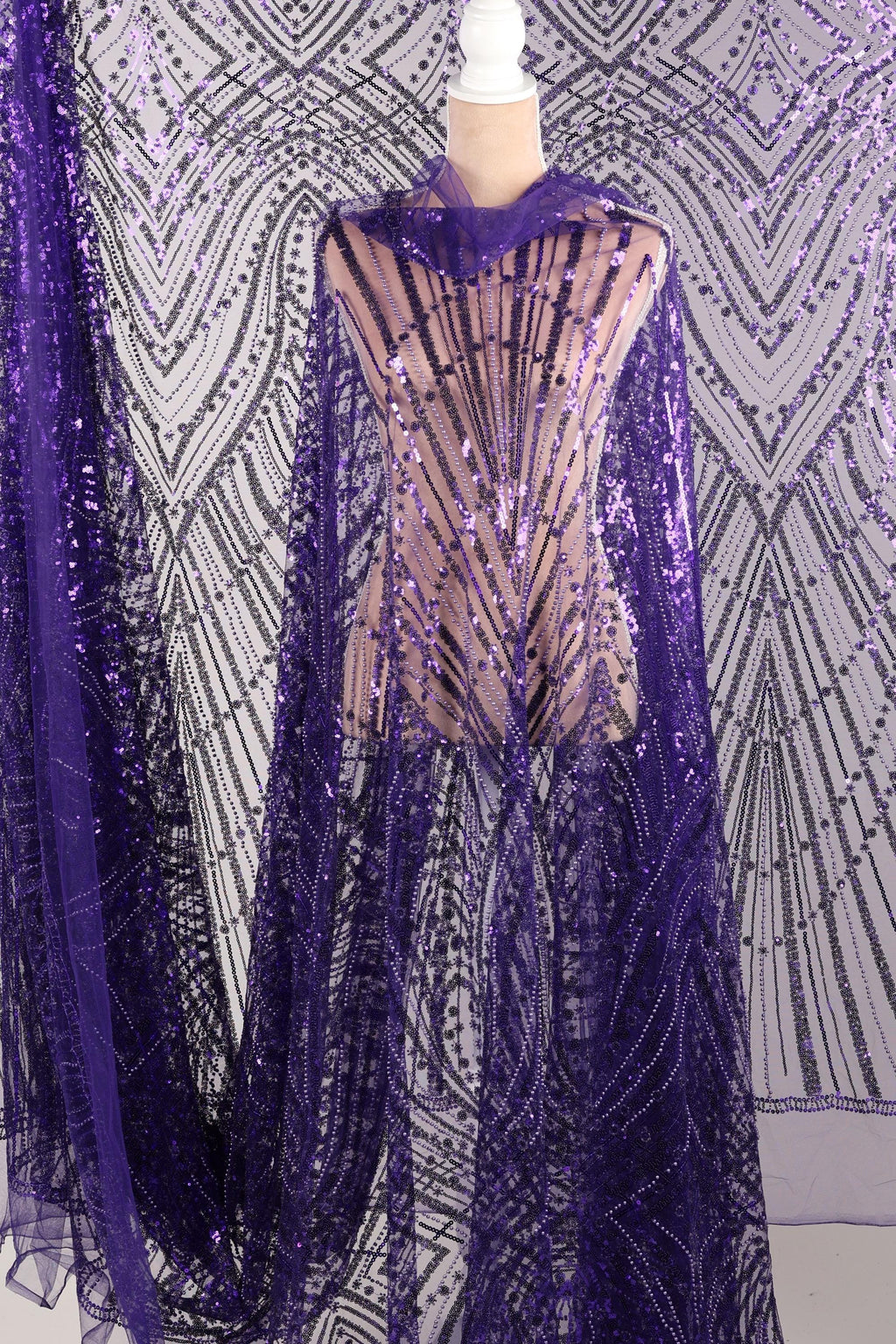 a mannequin wearing a purple sequin dress with a sheer overlay. The dress has a long, flowing cape-like design with a pattern of purple and black sequins. The mannequin is positioned in front of a background with a similar pattern of purple and black sequins.