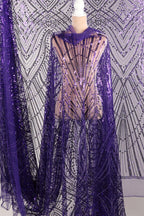 a mannequin wearing a purple sequin dress with a sheer overlay. The dress has a long, flowing cape-like design with a pattern of purple and black sequins. The mannequin is positioned in front of a background with a similar pattern of purple and black sequins.