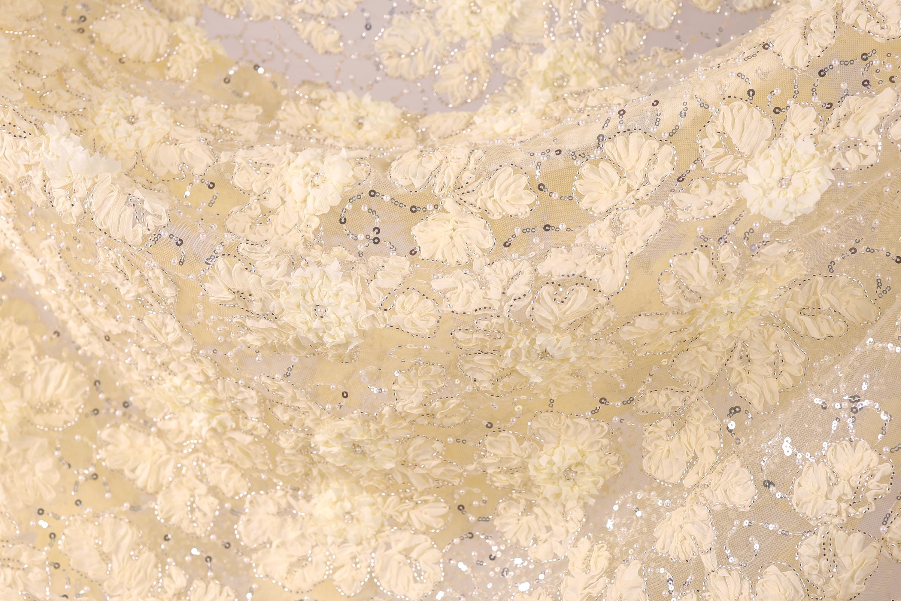 The image is a close-up of a fabric with a floral pattern, featuring white and beige colors. The fabric appears to be made of a soft, delicate material, possibly lace or chiffon. The pattern consists of numerous small white flowers or leaves, interspersed with silver or metallic accents, creating a visually appealing and intricate design.