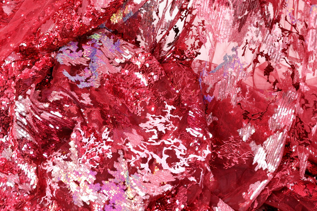 The image is a close-up of a fabric with a red and pink color scheme, featuring a variety of textures and patterns, including sequins and lace-like elements.