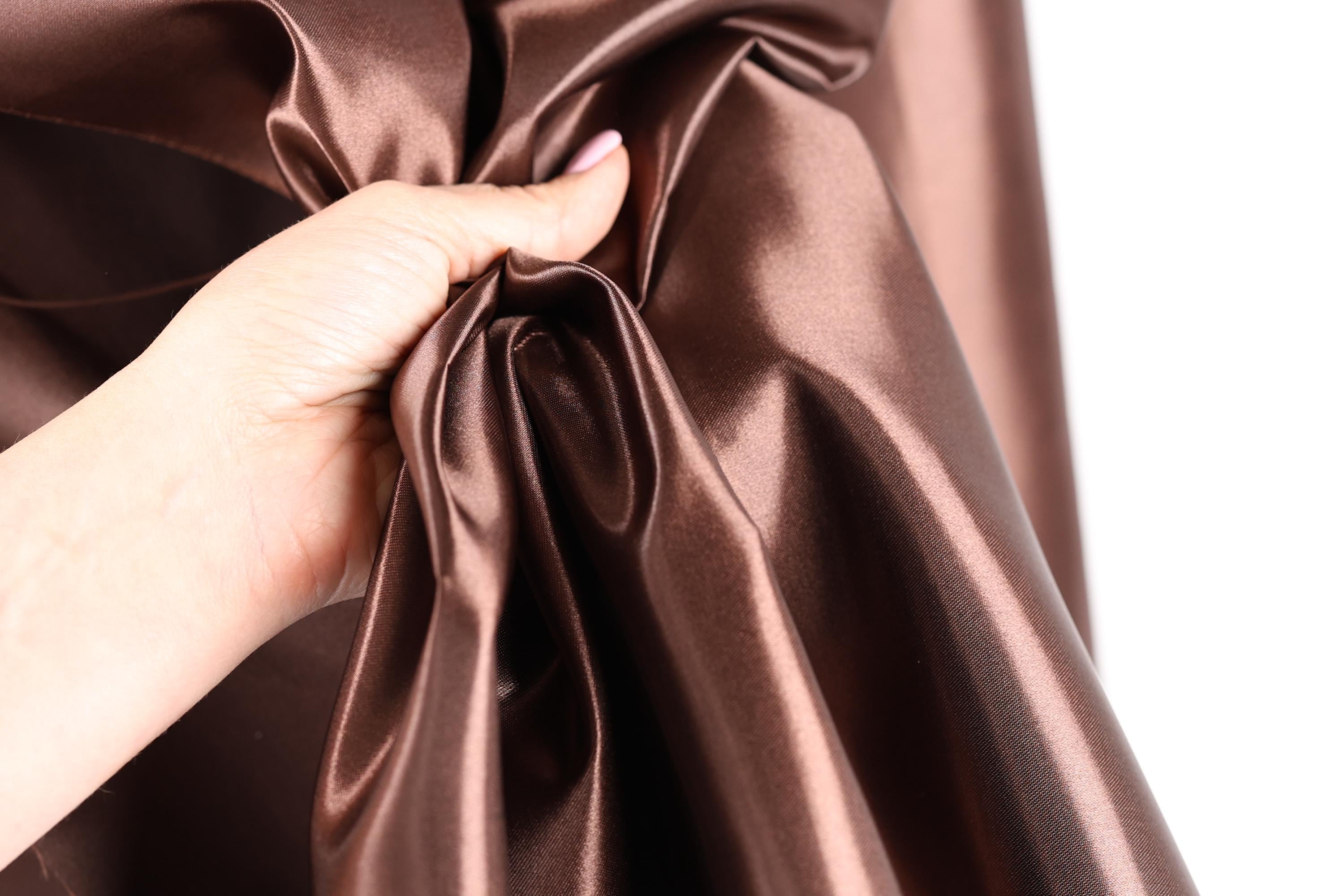 A hand is holding a piece of shiny brown satin fabric, which appears to be a close-up view of the material.