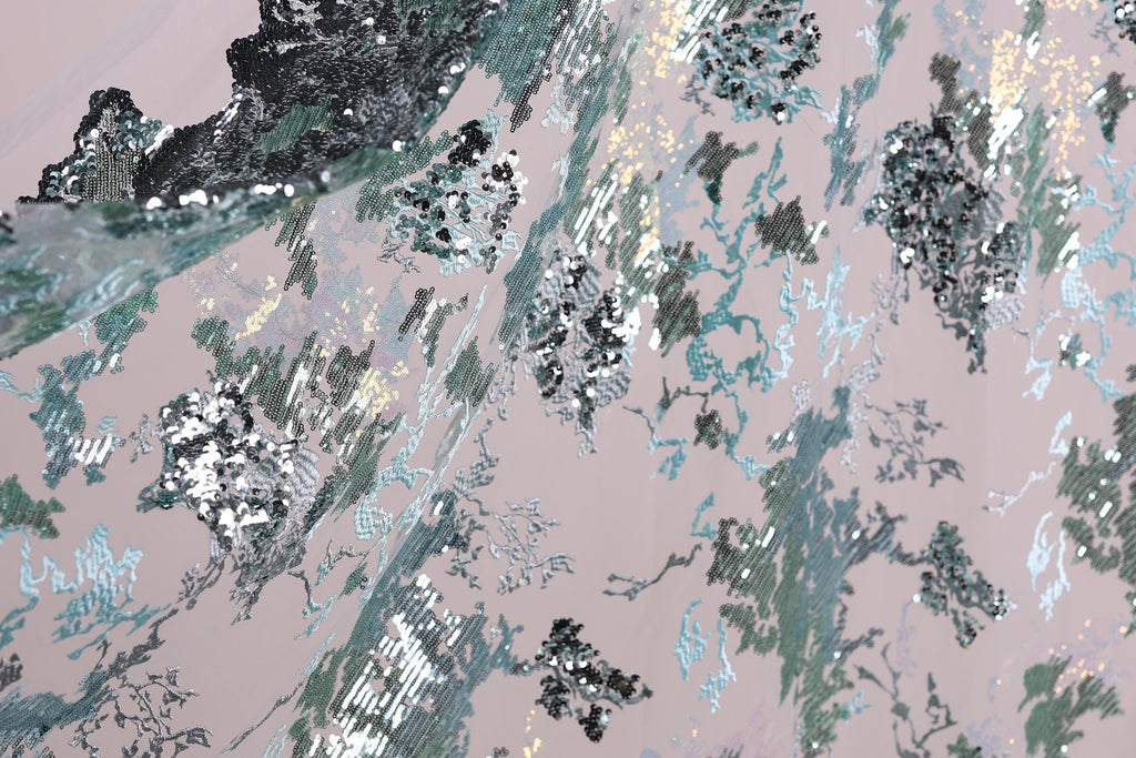 The image is a close-up view of a fabric with a pattern of sequins and glitter in shades of blue, green, and silver. The fabric appears to be a piece of clothing or textile, with the intricate design and shimmering sequins creating a visually striking and textured appearance.