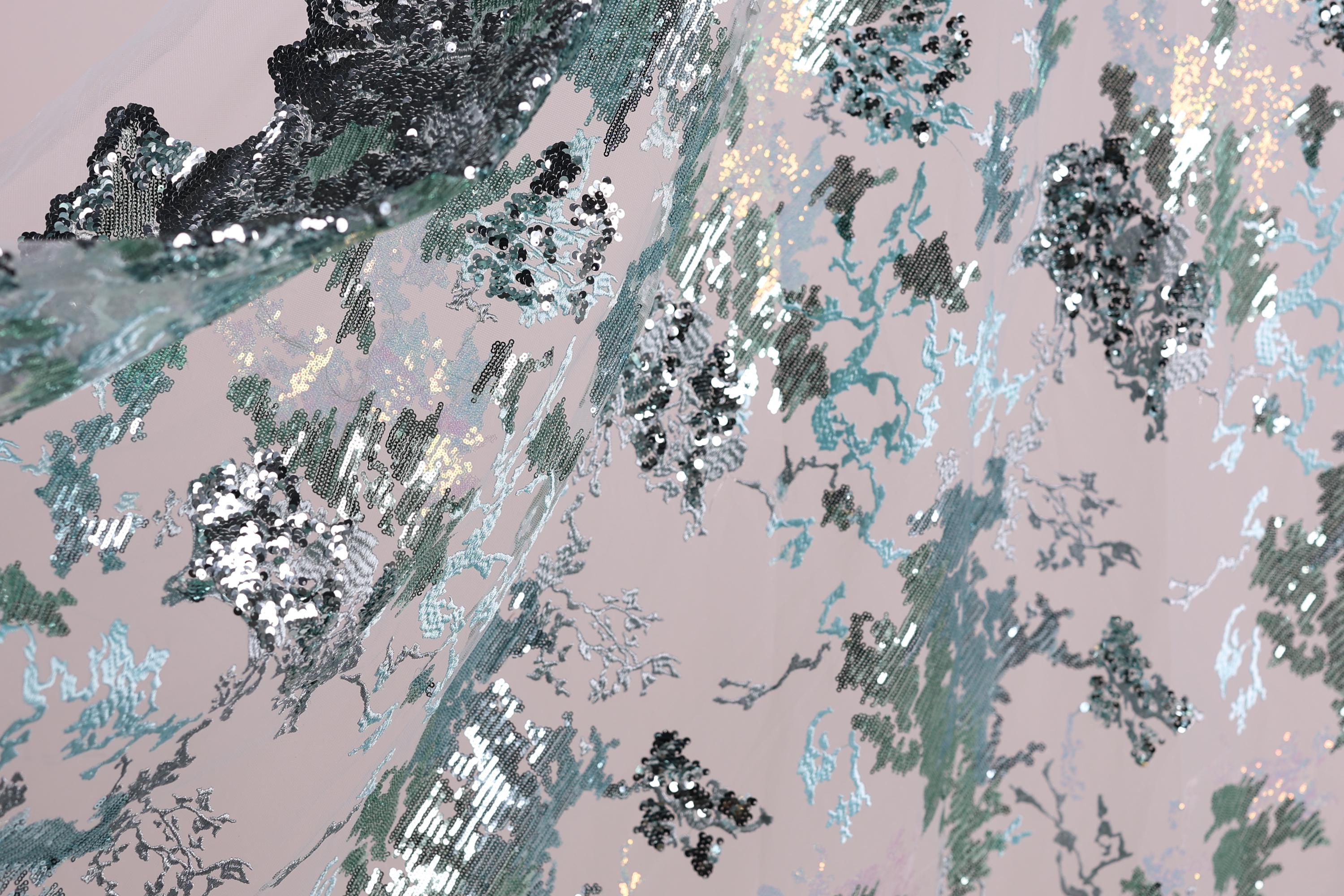 The image is a close-up view of a fabric with a pattern of sequins and glitter in shades of blue, green, and silver. The fabric appears to be a piece of clothing or textile, with the intricate design and shimmering sequins creating a visually striking and textured appearance.